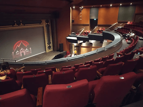 Interior view of a Balgandharva Rangmandir auditorium with red seating, curved balcony, and a stage displaying an illuminated architectural graphic